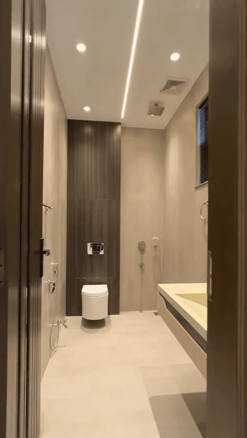 Modern minimalist bathroom with a wall-mounted toilet, dark wood accent wall, and linear ceiling lighting.